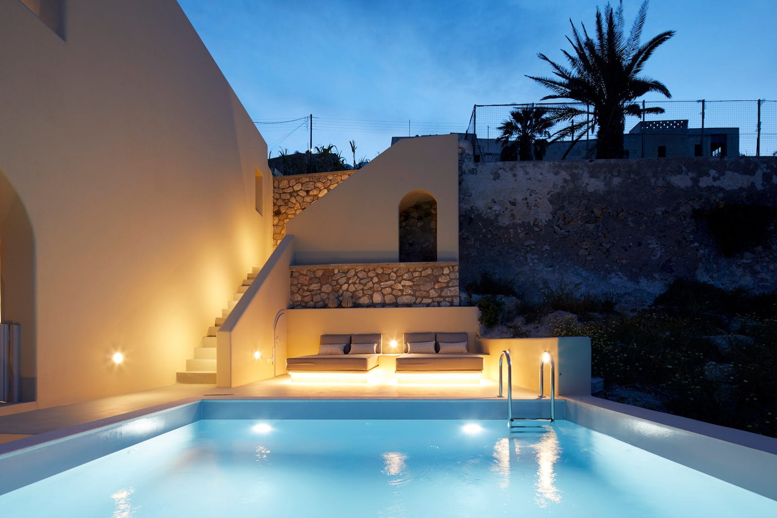 Illuminated swimming pool at twilight with cushioned sunbeds set against traditional Cycladic stone terracing